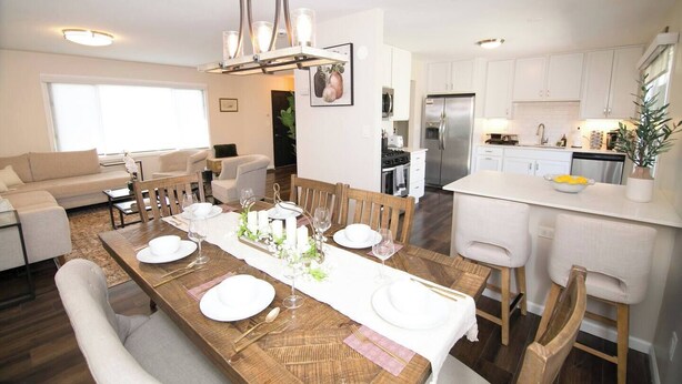 Un coin repas avec une table en bois dressée pour quatre personnes, une cuisine avec des meubles blancs et un salon avec un canapé et des fauteuils.