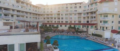 A hotel with a swimming pool surrounded by lounge chairs and umbrellas, multiple balconies, and a covered seating area.