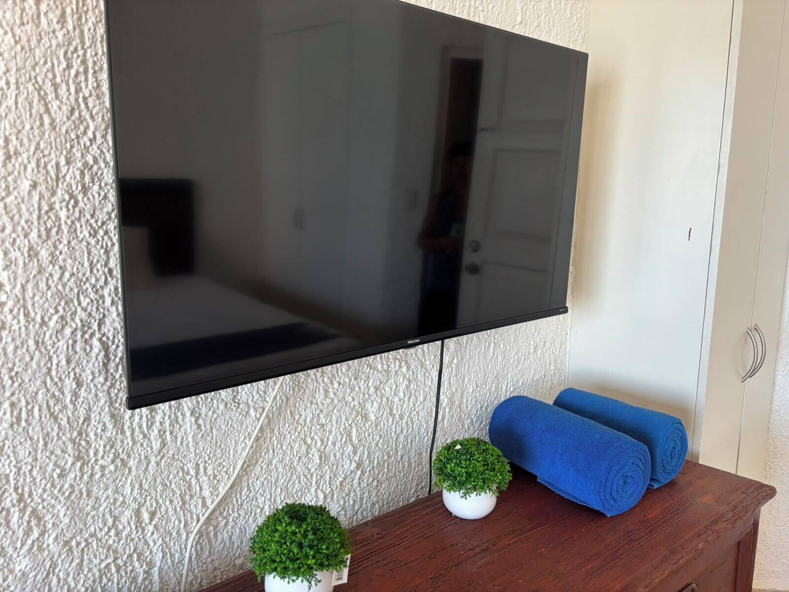 A flat-screen TV mounted on a textured wall, a small potted plant, and a wooden table with rolled-up blue towels.