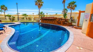 Piscina com borda azulejada, cercada por palmeiras e um edifício com fachada amarela.
