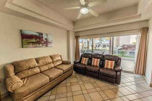 Ein Wohnzimmer mit braunem Sofa, einem braunen Ledersofa mit gestreiften Kissen, einem Deckenventilator und einem großen Fenster mit Blick ins Freie.