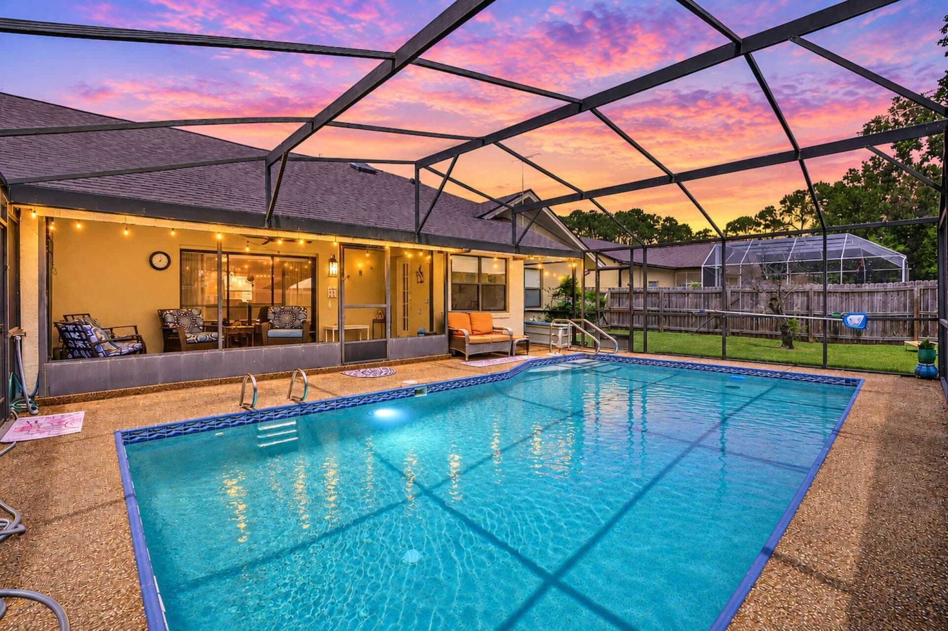 Ein Schwimmbad mit überdachter Terrasse, einem eingezäunten Garten und einem Sonnenuntergangshimmel.