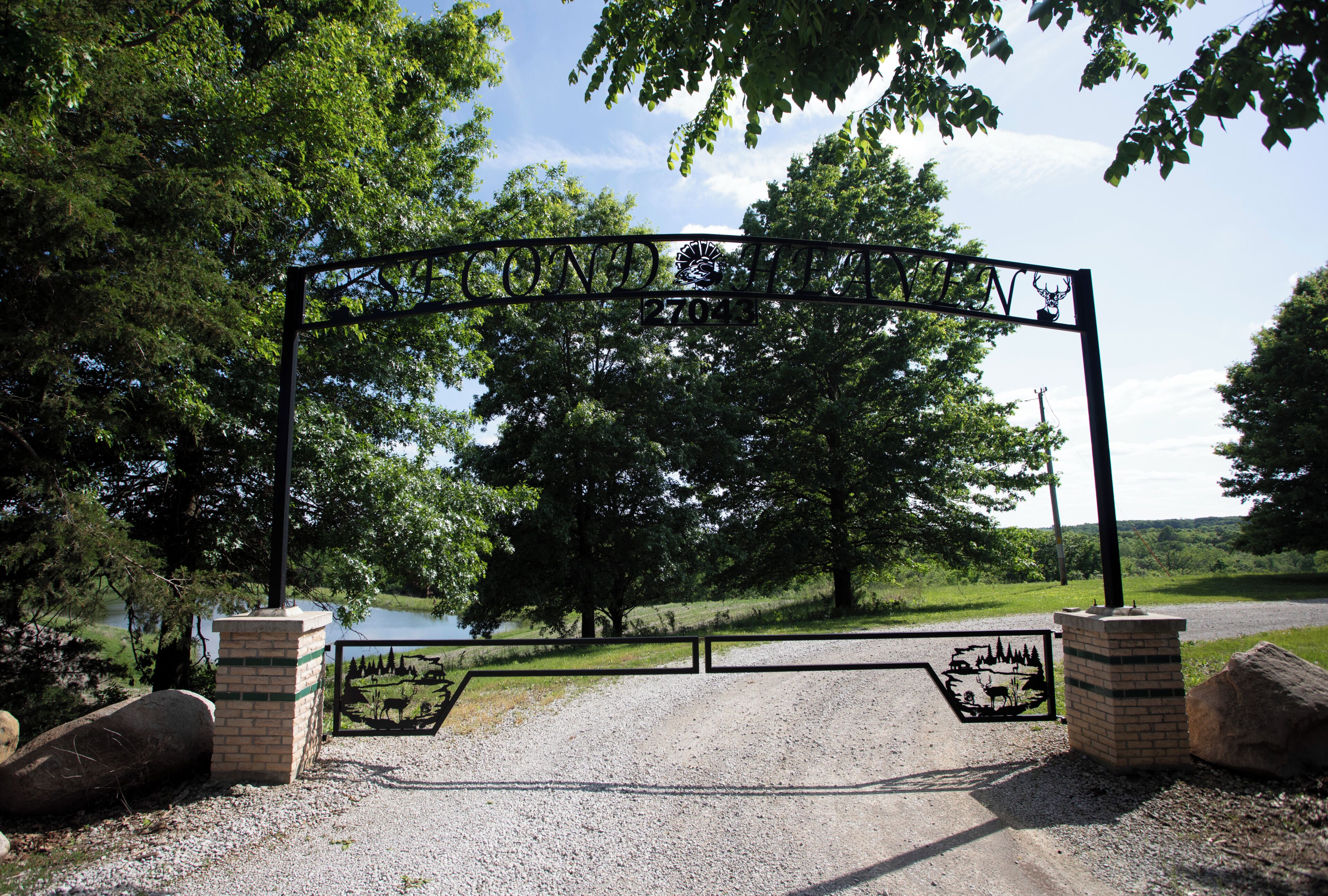 A metal gate with a deer silhouette, a brick structure, and a gravel path leading to a body of water.