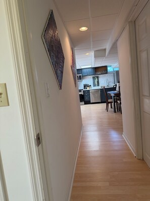 A hallway leading to a kitchen with wooden flooring, white walls, and a ceiling with recessed lighting.