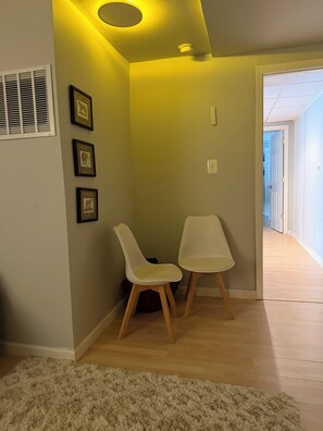 A room with a white chair, a white table, and a fluffy rug. There are framed pictures on the wall and a vent on the wall.