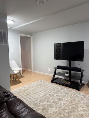 Un salon avec un téléviseur à écran plat posé sur un meuble noir, une chaise blanche, un tapis blanc et un plancher en bois.