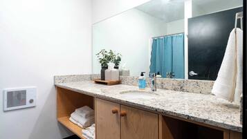 A bathroom with a granite countertop, a sink, a mirror, a towel rack, a shelf with folded towels, a potted plant, and a shower curtain.