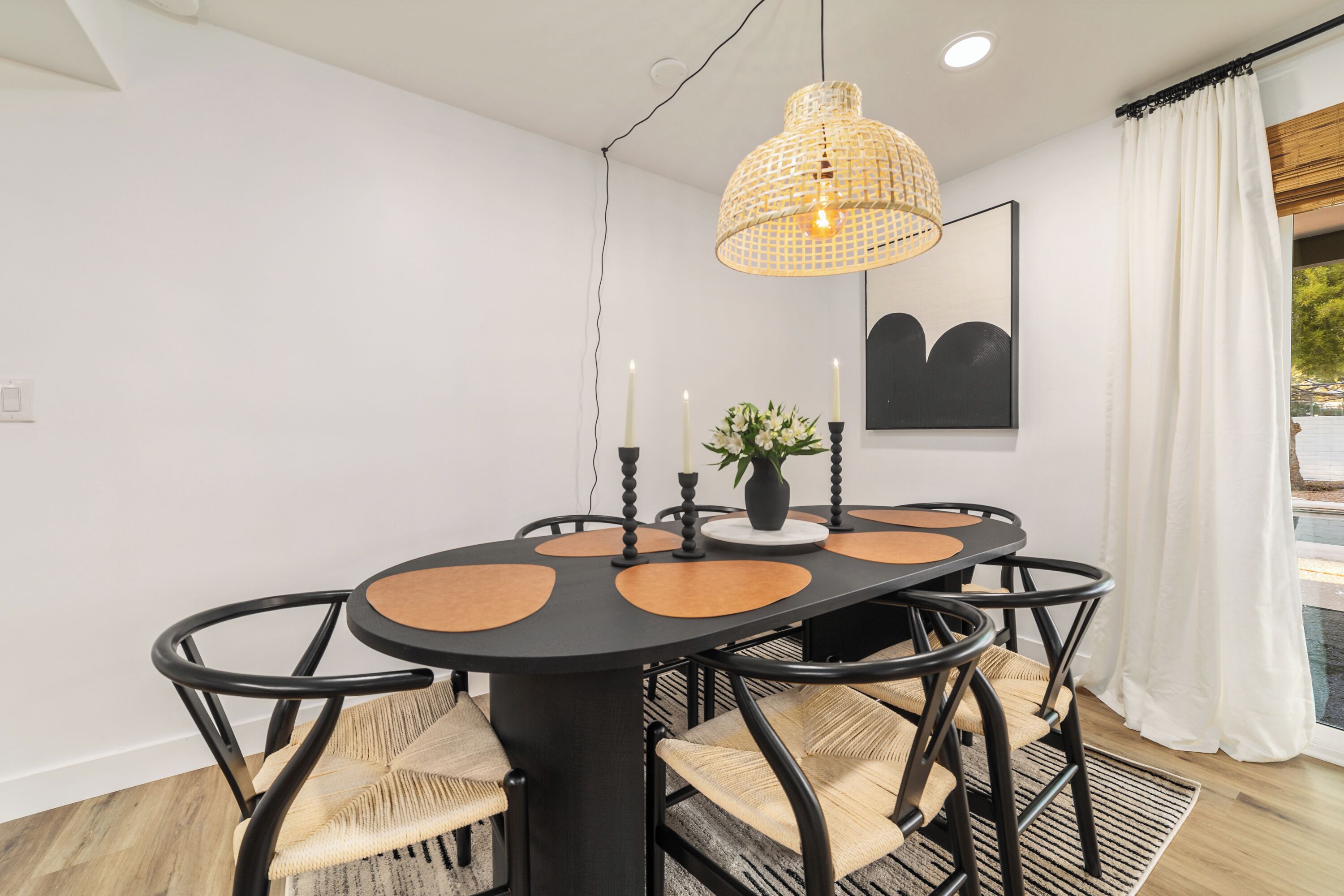 A dining area with a round table, black chairs, a woven pendant light, candles, and a framed artwork on the wall.