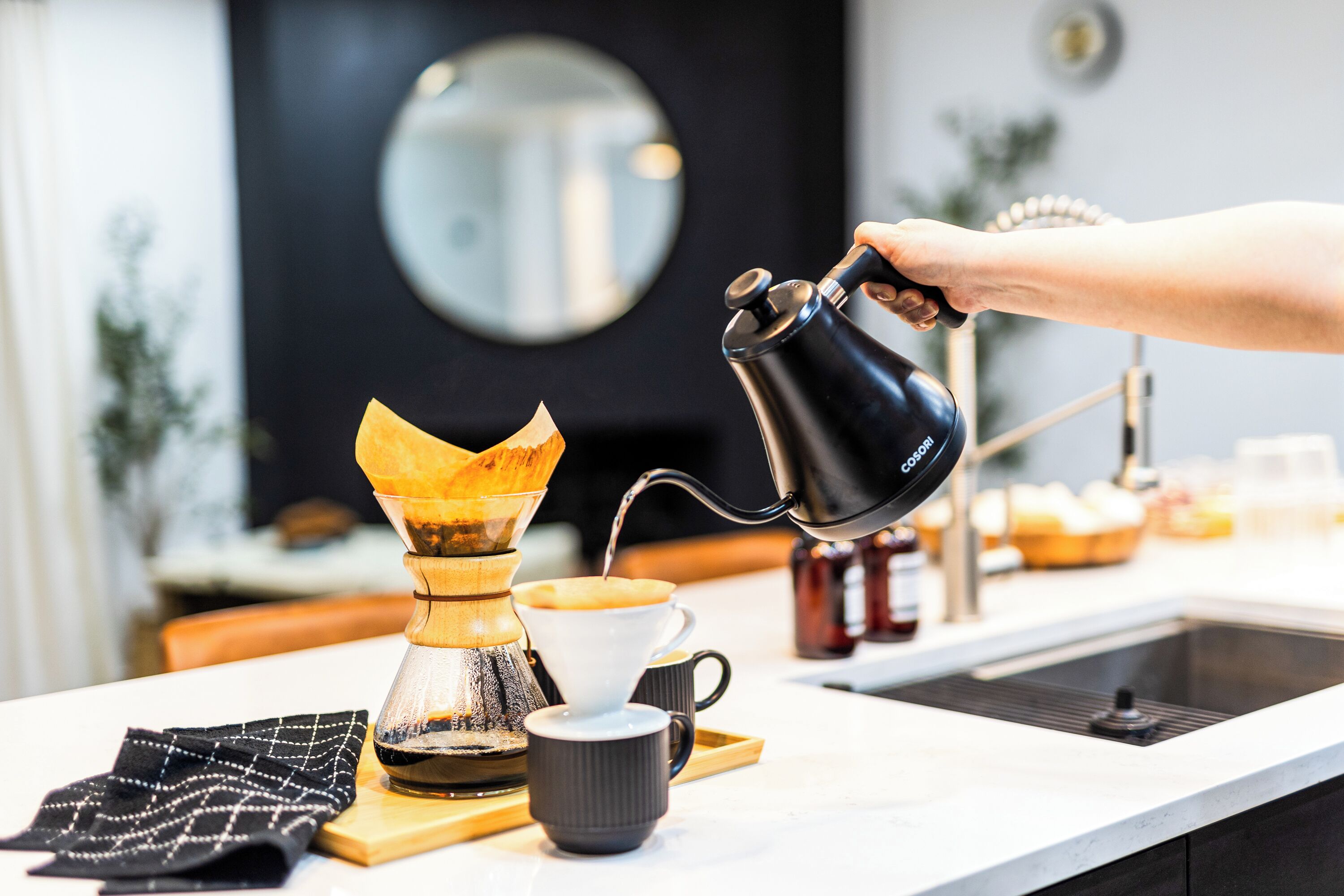 Eine Hand gießt Kaffee in eine Tasse, ein Kaffeemühle und eine Kaffeemaschine stehen auf einer Küchenarbeitsplatte.