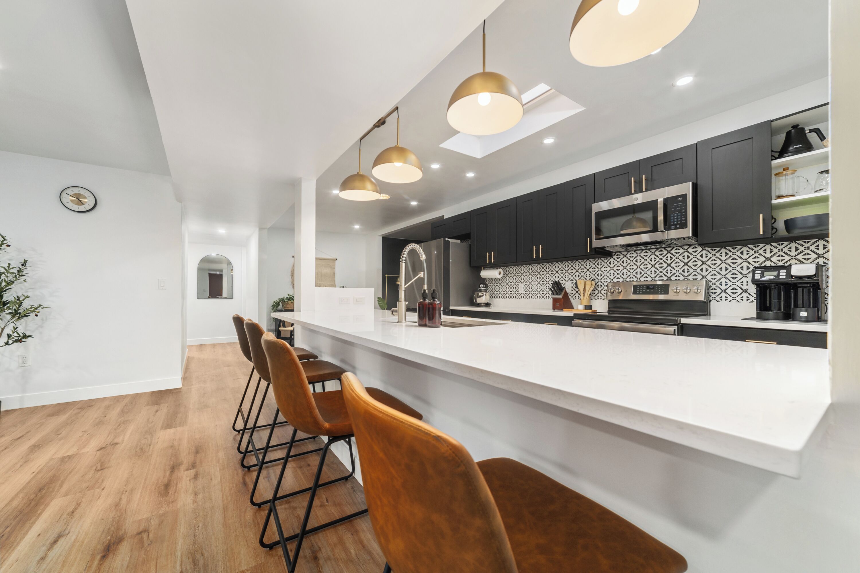 Une cuisine moderne avec un plan de travail blanc, des meubles de couleur sombre et un micro-ondes encastré. On y trouve trois lampes suspendues, un évier en acier inoxydable et un coin bar avec des tabourets.