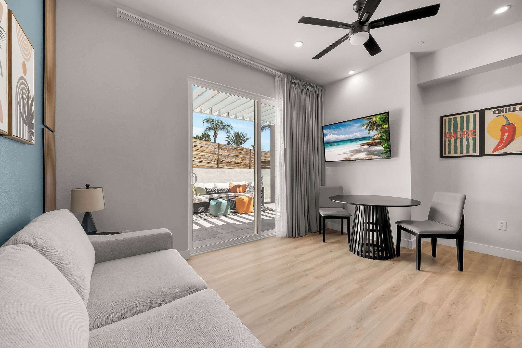 Ein modernes Wohnzimmer mit einer Couch, einem Esstisch mit Stühlen, einem Deckenventilator und einem großen Fenster mit Blick auf einen Strand.