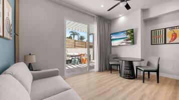 A modern living room with a sofa, a dining table with chairs, a ceiling fan, and a large window with a view of a beach.