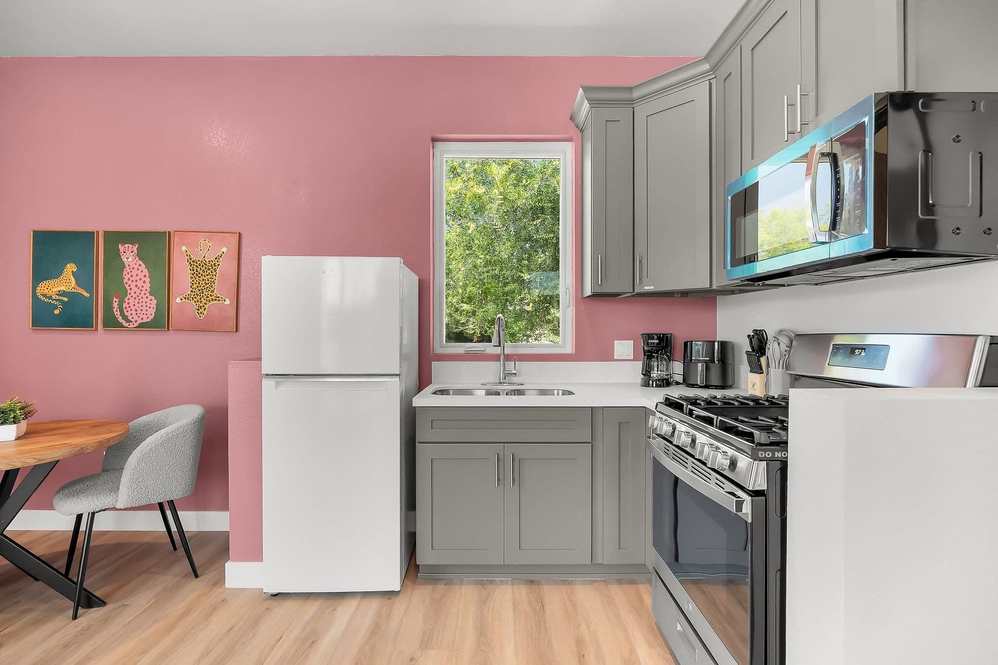 A modern kitchen with grey cabinets, a white refrigerator, a microwave, and a stove. There is a window with a view of trees, and a table with a chair.