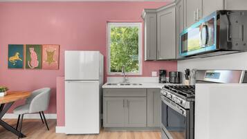 A modern kitchen with grey cabinets, a white refrigerator, a microwave, and a stove. There is a window with a view of trees, and a table with a chair.