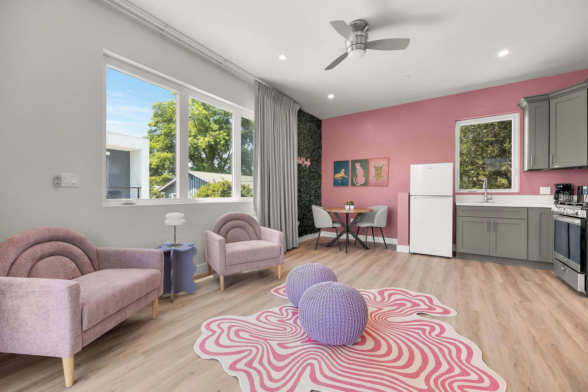 A modern apartment with a living room, dining area, and kitchen. There is a ceiling fan, large windows, and a view of the outside.