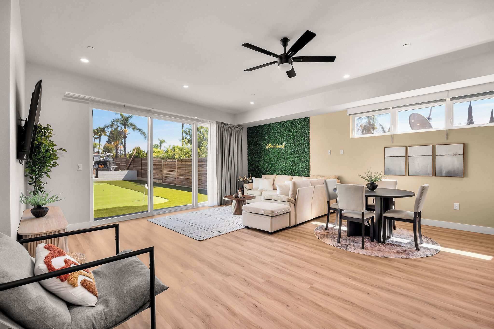 A modern living room with a flat-screen TV, a ceiling fan, a green wall, and a dining area with a table and chairs.