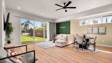 A modern living room with a flat-screen TV, a ceiling fan, a green wall, and a dining area with a table and chairs.