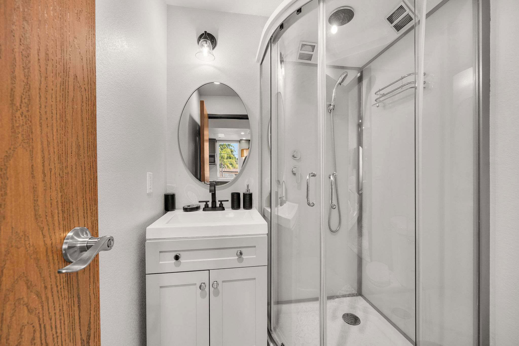 A bathroom with a wooden door, a white vanity with a mirror, and a glass-enclosed shower.