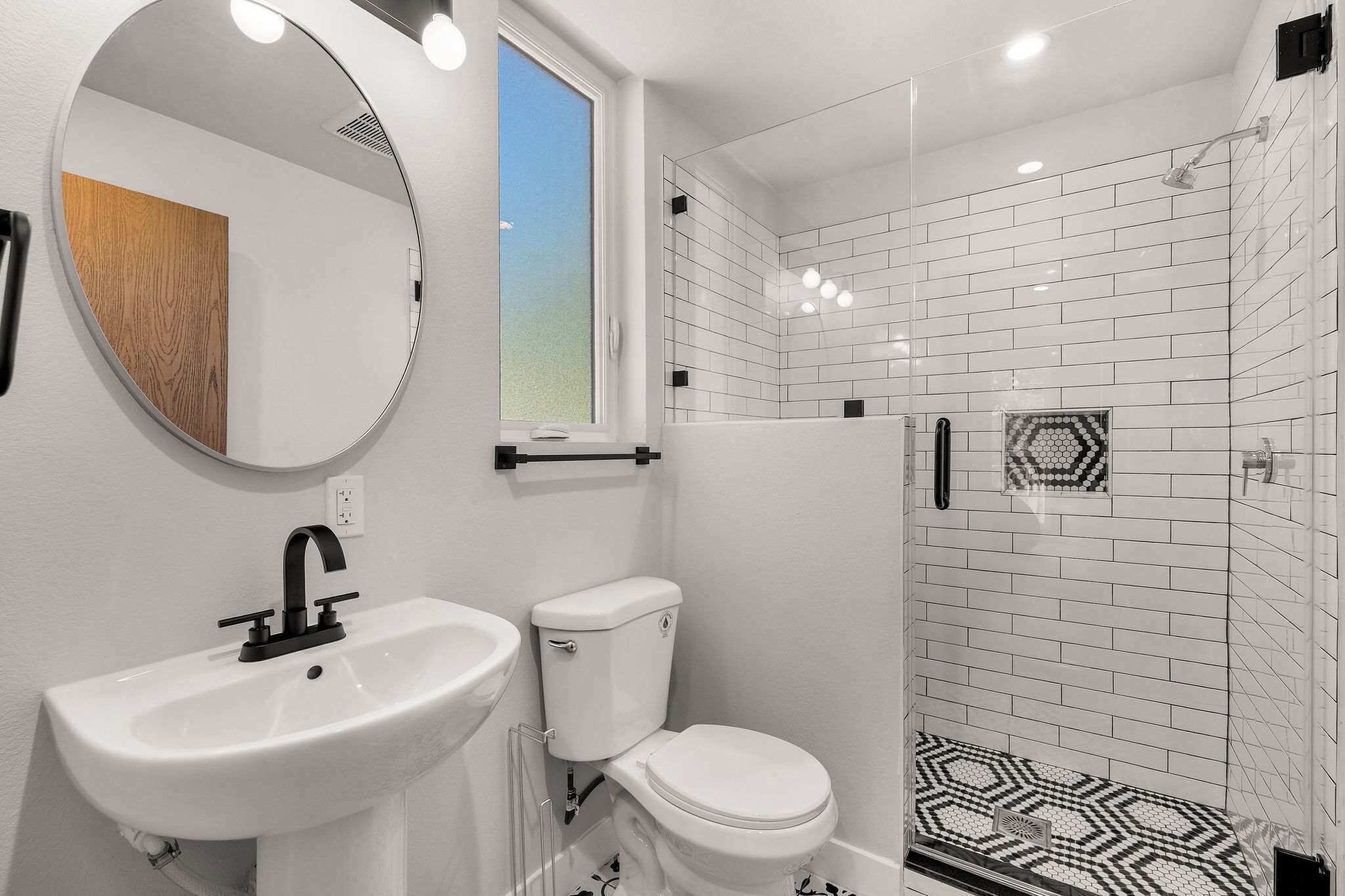 Un baño con lavamanos, inodoro y ducha blancos. Hay una ventana sobre el lavamanos y un espejo en la pared.