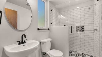 Un baño con lavamanos, inodoro y ducha blancos. Hay una ventana sobre el lavamanos y un espejo en la pared.