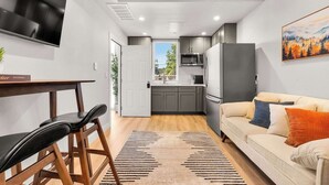 A modern kitchen with grey cabinets, a white refrigerator, and a microwave. There is a dining area with a wooden table and chairs. A beige sofa with colorful cushions is in the living area.