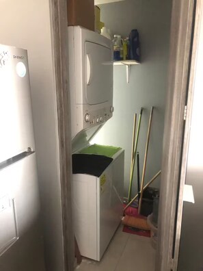 Een compacte keuken met een koelkast, magnetron en wasmachine. Er staan schoonmaakspullen en een bezem in de hoek.