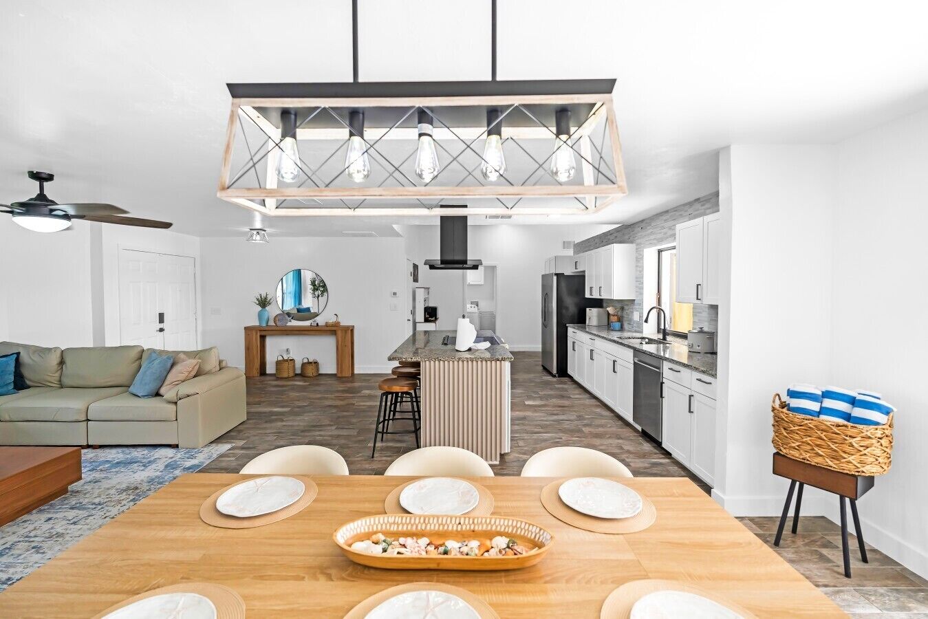 A modern kitchen with a central island, a dining area with a wooden table, and a living room with a sofa and a ceiling fan.
