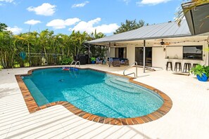 A swimming pool with a wooden deck, a covered seating area, and a building with a metal roof.