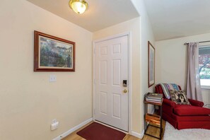 Ein Zimmer mit weißer Tür, einem gerahmten Gemälde, einem roten Sessel, einem kleinen Tisch mit Büchern und einem Fenster mit Vorhängen.