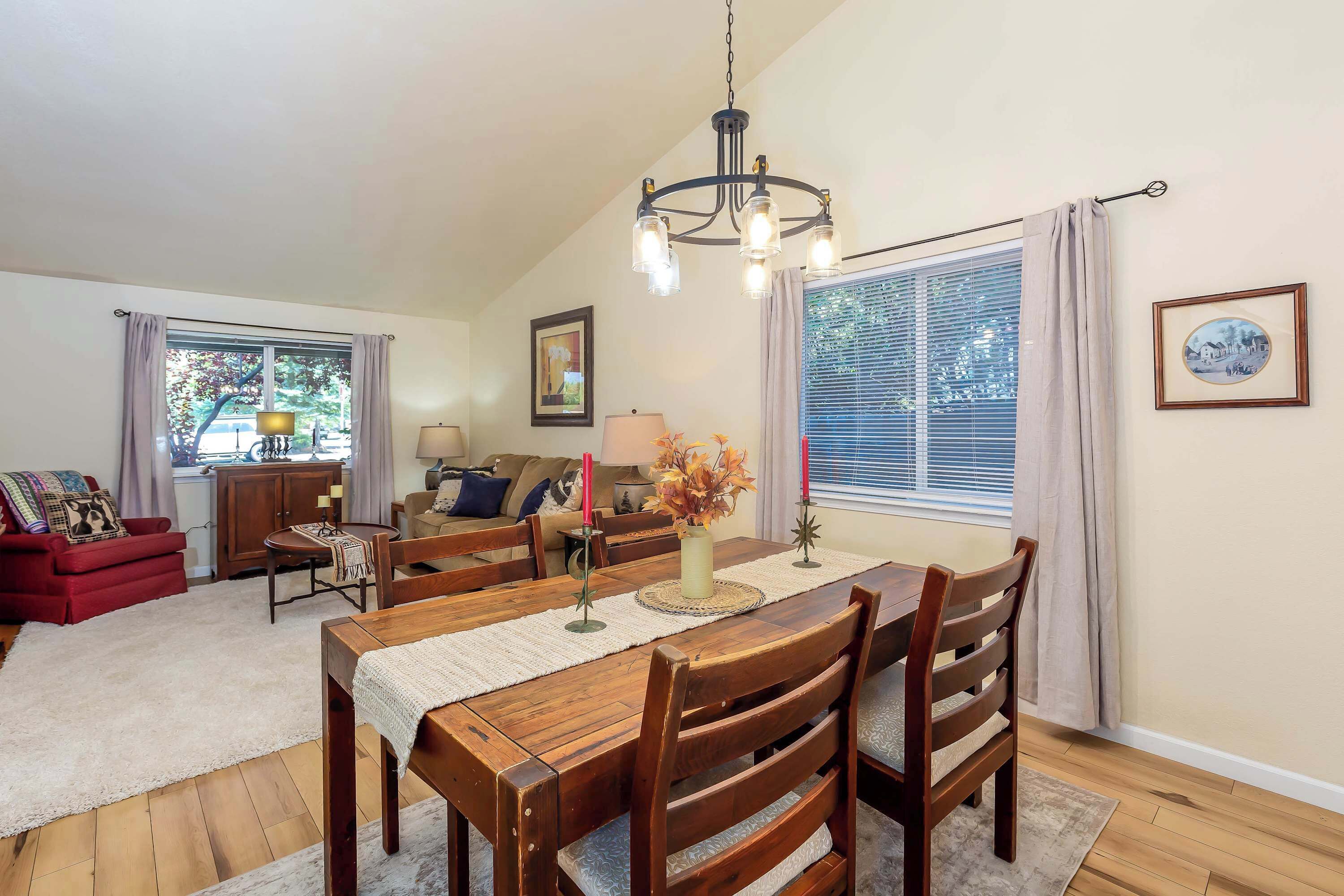 A dining area with a wooden table and chairs, a chandelier, and a window with blinds.