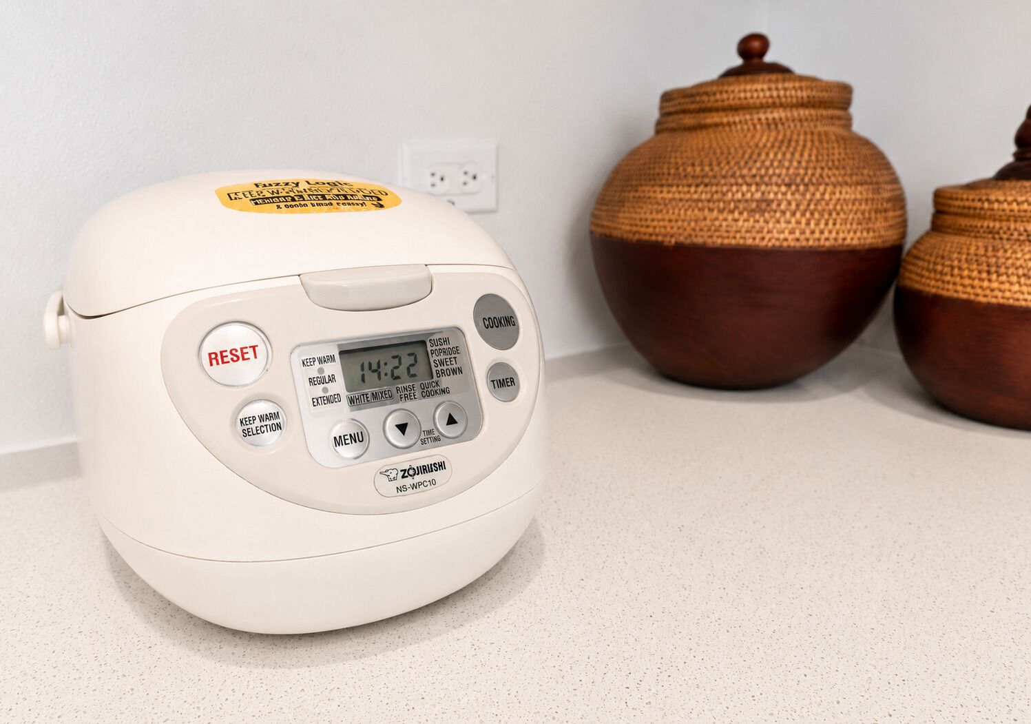 Una olla arrocera blanca con pantalla digital, botones para configuraciones y la etiqueta de la marca.