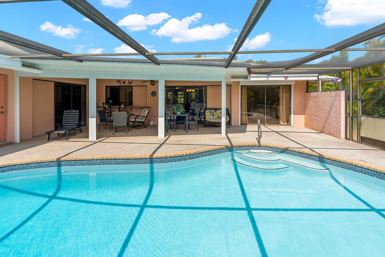 En swimmingpool med flisekant, en overdækket terrasse med udendørs møbler og en klar blå himmel.