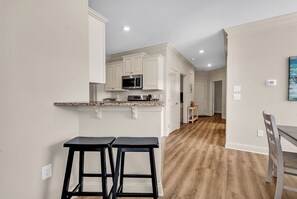 Une cuisine moderne avec des meubles blancs, un four à micro-ondes et un plan de travail. On y trouve deux tabourets noirs et une table à manger en bois avec une chaise. Le sol est en bois et les murs sont peints dans une couleur claire.