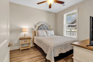 Une chambre à coucher comprenant un lit, une table de chevet, une lampe, un ventilateur de plafond, une fenêtre avec des stores et un téléviseur.