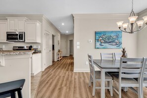 A modern kitchen with white cabinetry, a stainless steel microwave, and a dining area with a wooden table and chairs. - Sunnyside Village-3A condo near Marshwalk with pool (Murrells Inlet)
