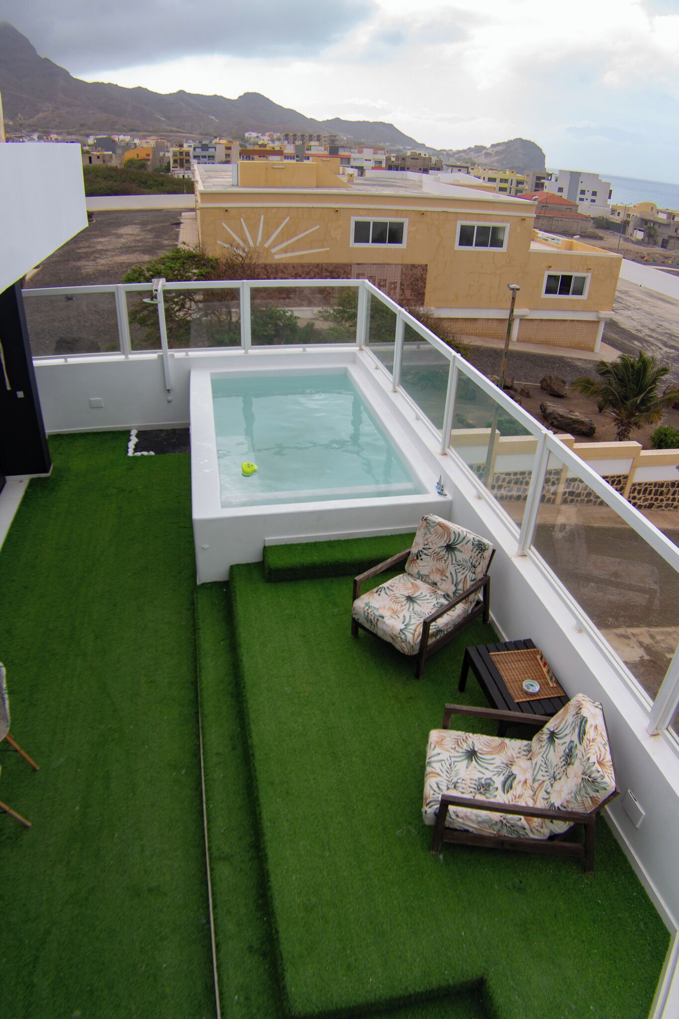 A rooftop terrace with a pool, two lounge chairs, and a view of a coastal town.