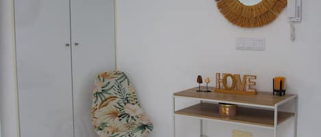 A bathroom with a white vanity, a mirror with a woven frame, a chair with a tropical print, and a small rug on the floor.