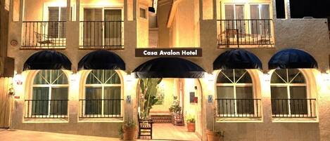 A hotel with a sign reading 'Casa Avalon Hotel,' featuring a central entrance with a covered walkway, potted plants, and a balcony with a railing.