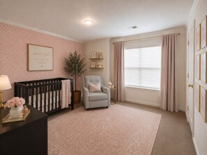 Ein Babyzimmer mit einem Kinderbett, einem Stuhl, einem Fenster mit Vorhängen, einer Wand mit einem Bild und einem kleinen Tisch mit einer Lampe.