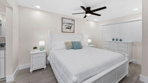 Un dormitorio con una cama blanca, dos mesitas de noche con lámparas, un ventilador de techo y un cuadro en la pared.