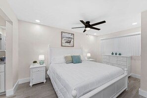 Ein Schlafzimmer mit einem weißen Bett, zwei Nachttischen mit Lampen, einem Deckenventilator und einem Bild an der Wand.