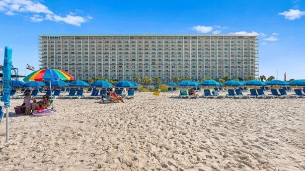 Una playa con hileras de sombrillas azules y sillas de descanso, un gran edificio de hotel al fondo, y un cielo azul despejado.