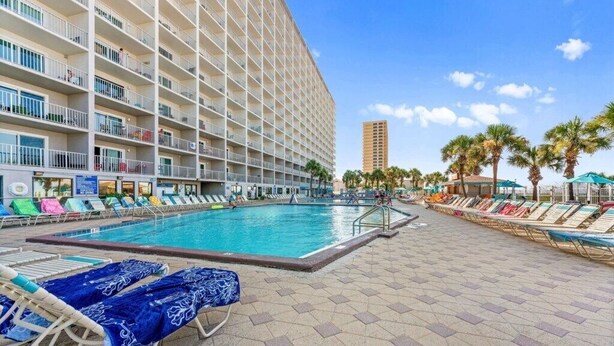 Zona de piscina de hotel con sillones de descanso, una piscina y un edificio alto al fondo.