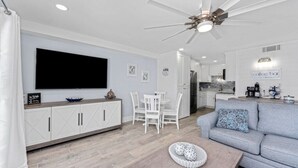Una sala de estar moderna con televisor de pantalla plana, muebles blancos, ventilador de techo y una cocina al fondo.