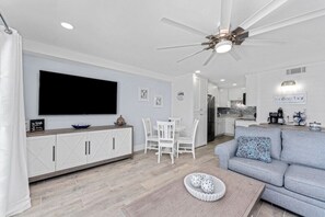 Ein modernes Wohnzimmer mit Flachbildfernseher, weißen Möbeln, Deckenventilator und einer Küche im Hintergrund.
