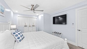 Un dormitorio con una cama, un ventilador de techo, un televisor montado en la pared y una puerta blanca.