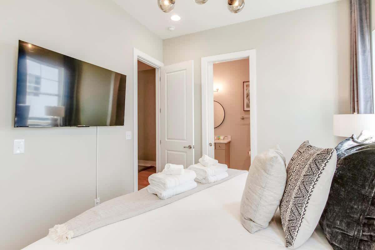 A hotel room with a bed, a flat-screen TV mounted on the wall, a bathroom visible through an open door, and a towel rack with towels.