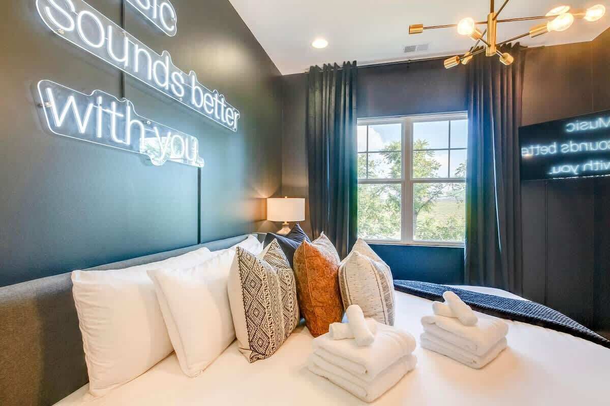 Une chambre d’hôtel avec un lit, des oreillers, une table de chevet avec une lampe, un mur avec un texte illuminé et une fenêtre avec des rideaux.