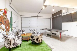 Ein Zimmer mit Tischtennisplatte, zwei Stühle mit Kuhmuster, einem kleinen runden Tisch und einem Wandbild.