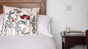 Ein Bett mit einem blumigen Kissen, einem Holz-Kopfteil, einem Nachttisch mit Buch und Kerze sowie ein Sessel.